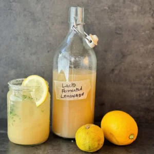 A photo of a glass bottle of fermented lemonade, a small glass of lemonade next to it and lemons