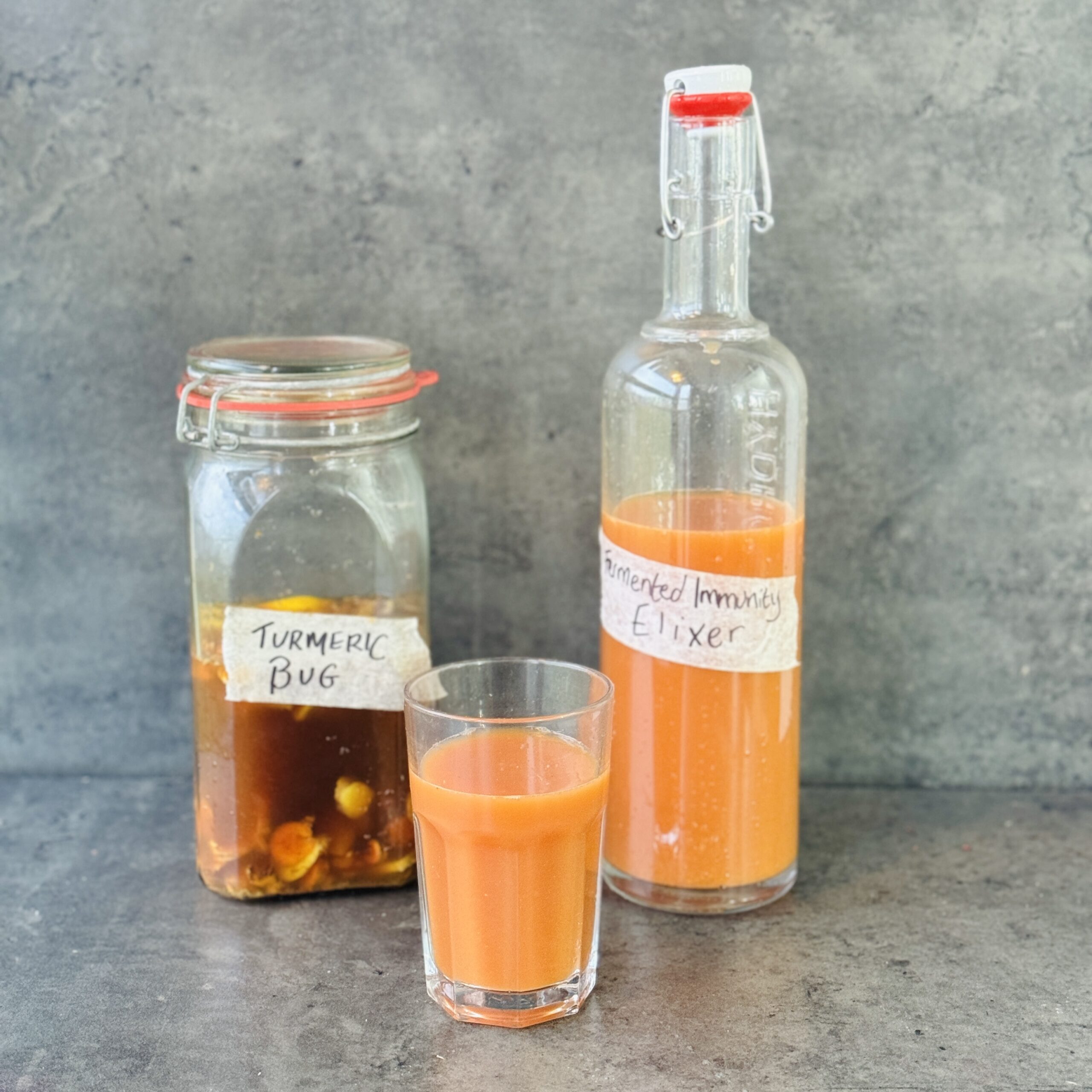 a photo of a bottle and glass of Fermented Immunity Elixir next to a jar labelled turmeric bug